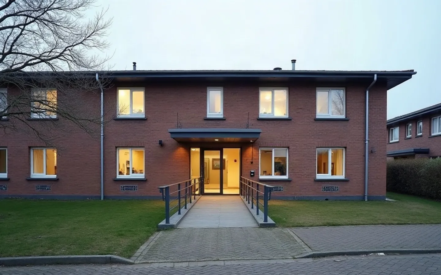 Laag zorggebouw van baksteen in de vroege ochtend, met brede ramen op de begane grond, een overdekte ingang met schuifdeur, een hellingbaan met leuning en een leeg voorplein.